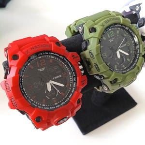 May include: Two digital watches, one red and one olive green, are displayed. The red watch features a black face with white hands and text, including "X NATION" and "12." The green watch has a similar design. Both watches are on a black stand.