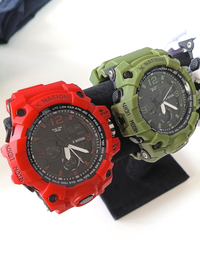 May include: Two digital watches, one red and one olive green, are displayed. The red watch features a black face with white hands and text, including "X NATION" and "12." The green watch has a similar design. Both watches are on a black stand.