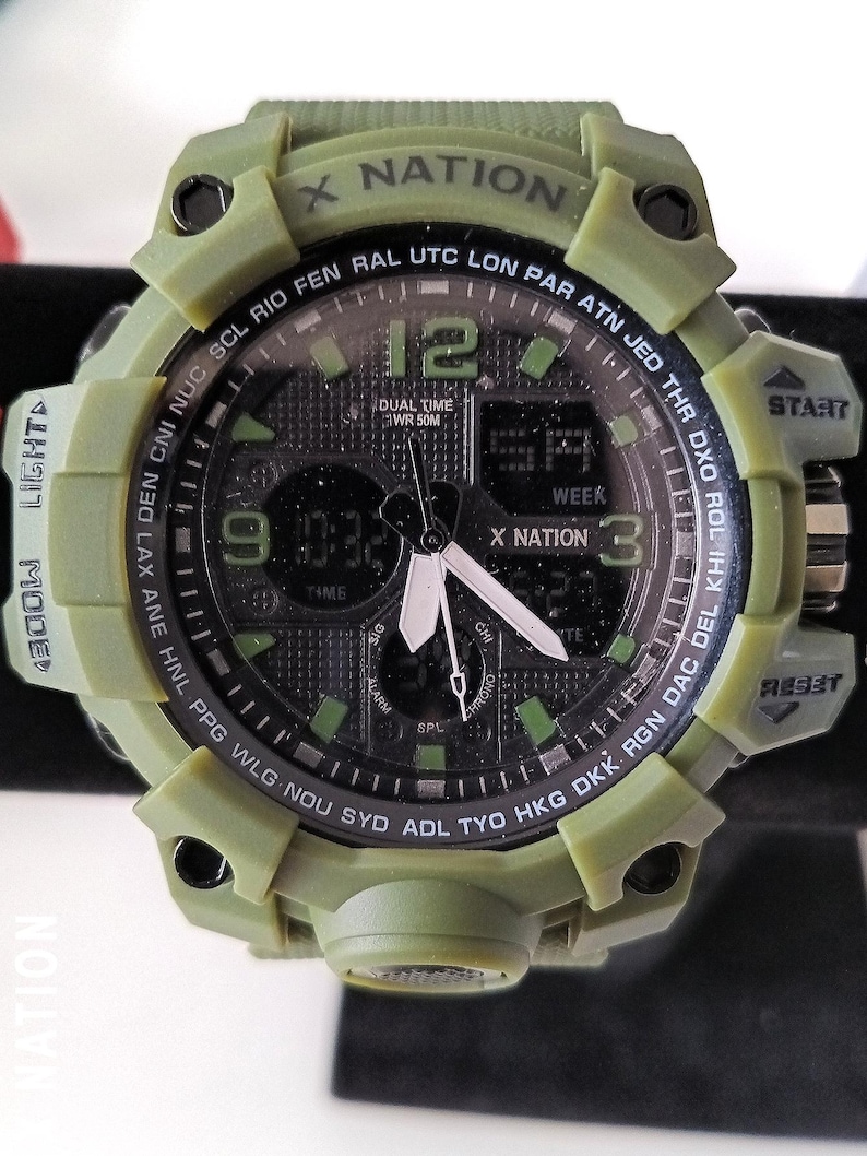 May include: An olive green digital watch with the text "X NATION" on the bezel. The watch face displays the time, day, and date. It has multiple buttons for various functions, including "START" and "RESET".