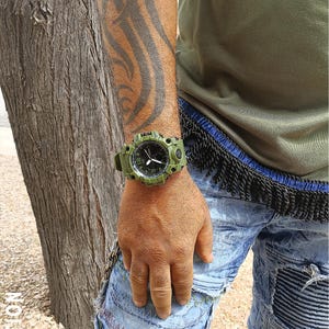 May include: An olive green digital watch with a black dial is worn on a wrist. The watch has a rugged design with a green strap and casing. The person is wearing a green shirt and blue jeans.