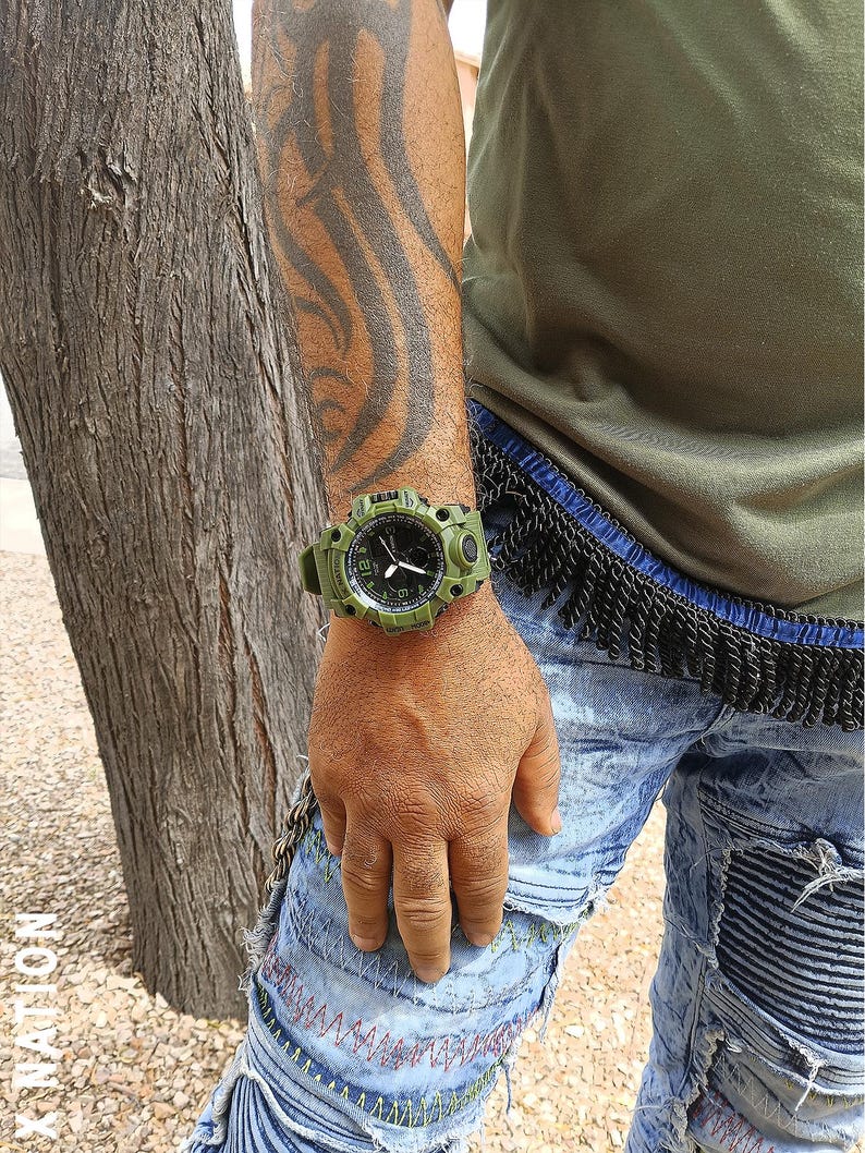May include: An olive green digital watch with a black dial is worn on a wrist. The watch has a rugged design with a green strap and casing. The person is wearing a green shirt and blue jeans.