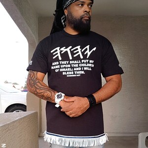 May include: Black t-shirt with white Hebrew text and the English quote "AND THEY SHALL PUT MY NAME UPON THE CHILDREN OF ISRAEL; AND I WILL BLESS THEM." The shirt has a fringed hem and is paired with blue jeans.