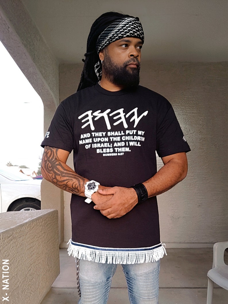 May include: Black t-shirt with white Hebrew text and the English quote "AND THEY SHALL PUT MY NAME UPON THE CHILDREN OF ISRAEL; AND I WILL BLESS THEM." The shirt has a fringed hem and is paired with blue jeans.