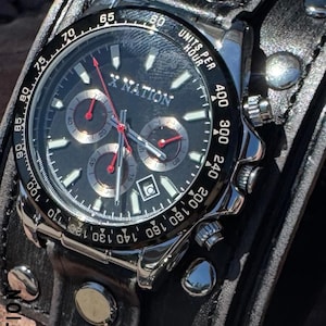 May include: A black and silver wristwatch with a black leather band. The watch face features the text "X NATION" and has three sub-dials with red hands. The band is wide and has silver rivets.