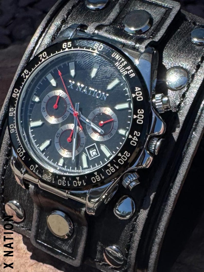 May include: A black and silver wristwatch with a black leather band. The watch face features the text "X NATION" and has three sub-dials with red hands. The band is wide and has silver rivets.