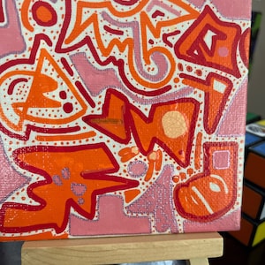 Puede incluir: Pintura abstracta sobre lienzo con formas y líneas naranjas y rosas. La pintura está en un pequeño caballete de madera.