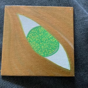 Puede incluir: Una pequeña pintura sobre lienzo de un ojo verde con texto amarillo dentro de la pupila. El ojo está sobre un fondo marrón.