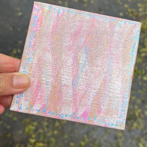 May include: Square canvas art with a textured surface. The artwork features a blend of pink, white, and blue hues, with subtle yellow accents. The edges are outlined with a delicate blue border, creating a soft, pastel aesthetic.