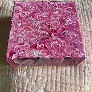 Puede incluir: Una pequeña pintura abstracta sobre lienzo en tonos rosa, blanco y azul. La pintura tiene un efecto de remolino y mármol.
