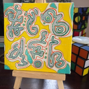 Puede incluir: Una pequeña pintura sobre lienzo amarillo con formas abstractas en gris, rojo y turquesa. La pintura está en un pequeño caballete de madera.