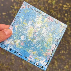 Puede incluir: Arte en lienzo cuadrado con un fondo azul claro y salpicaduras de pintura coloridas en blanco, amarillo y rosa. Los bordes están delineados con texto negro. La obra de arte está sostenida por una mano.
