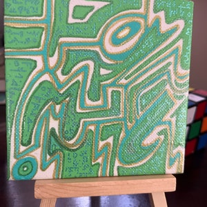 Puede incluir: Una pequeña pintura sobre lienzo verde y dorado con formas y líneas abstractas. La pintura está en un caballete de madera.