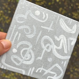 May include: A small, square canvas with a gray background and white abstract designs. The white designs include lines, swirls, and dots. The canvas is held by a person's hand.