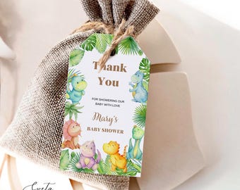 May include: A burlap drawstring favor bag with a rectangular paper tag attached by twine. The tag features green tropical leaves, colorful cartoon dinosaurs, and the text: Thank You, FOR SHOWERING OUR BABY WITH LOVE, Mary's BABY SHOWER.