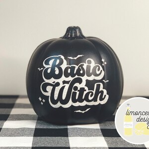 May include: Black pumpkin with the words "Basic Witch" in white glitter lettering. The pumpkin is sitting on a black and white checkered tablecloth.