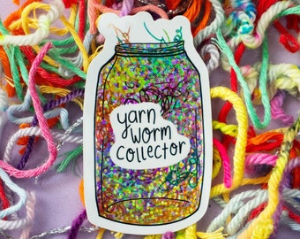 May include: A white sticker with a glitter-filled jar and the text "yarn worm collector". The sticker is surrounded by colorful yarn scraps.