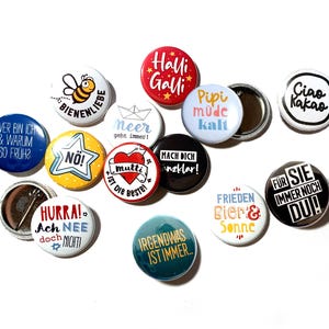 Könnte beinhalten: Eine Sammlung runder Ansteckbuttons mit verschiedenen bunten Motiven und deutschen Texten wie Halli Galli, Bienenliebe, Ciao Kakao, Wer bin ich & warum so früh, Nö, Mutti ist die Beste, Mach dich glasklar, Hurra Ach nee doch nicht, Irgendwas ist immer, Frieden Bier & Sonne, Pipi müde kalt, Meer geht immer und Für sie immer noch du.