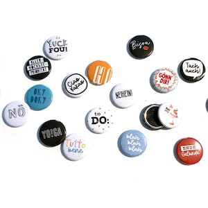 Peut inclure: Une collection de 16 boutons ronds colorés avec différentes phrases et designs. Les boutons présentent des phrases comme "Yuck Fou!" et "OK7 DOKY" en texte noir et blanc sur des fonds de couleurs variées.