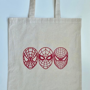 Puede incluir: Un bolso tote de lona beige con tres dise&ntilde;os de m&aacute;scaras rojas de Spider-Man. El bolso tiene dos asas y est&aacute; hecho de una tela resistente. Las m&aacute;scaras de Spider-Man est&aacute;n en fila.