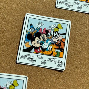 K&ouml;nnte beinhalten: Ein quadratischer Aufkleber im Polaroid-Stil mit Mickey Maus, Minnie Maus, Donald Duck, Daisy Duck, Goofy und Pluto. Unter den Charakteren sind die gedruckten Unterschriften von Mickey Maus, Minnie Maus, Pluto, Goofy, Donald Duck und Daisy Duck zu sehen.