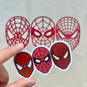 Puede incluir: Tres pegatinas de la cara de Spider-Man en rojo y blanco, sujetas por una mano. Las pegatinas presentan diferentes dise&ntilde;os de la m&aacute;scara del superh&eacute;roe. Tambi&eacute;n se ven tres contornos rojos de la m&aacute;scara en el fondo.