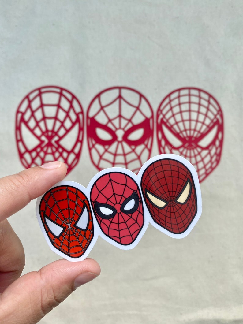Puede incluir: Tres pegatinas de la cara de Spider-Man en rojo y blanco, sujetas por una mano. Las pegatinas presentan diferentes dise&ntilde;os de la m&aacute;scara del superh&eacute;roe. Tambi&eacute;n se ven tres contornos rojos de la m&aacute;scara en el fondo.