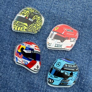 Puede incluir: Cuatro pegatinas en forma de casco de Fórmula 1. Las pegatinas presentan varios diseños, incluyendo remolinos amarillos y negros, rojo y blanco con el número 76, un diseño rojo y azul y un diseño azul claro con texto.