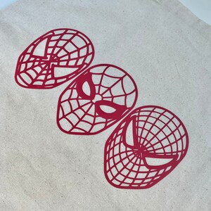 Puede incluir: Un bolso tote de lona beige con tres dise&ntilde;os de m&aacute;scaras rojas de Spider-Man. Las m&aacute;scaras presentan diferentes estilos, con patrones de telara&ntilde;a y formas de ojos. El bolso tiene una forma rectangular simple con dos asas.