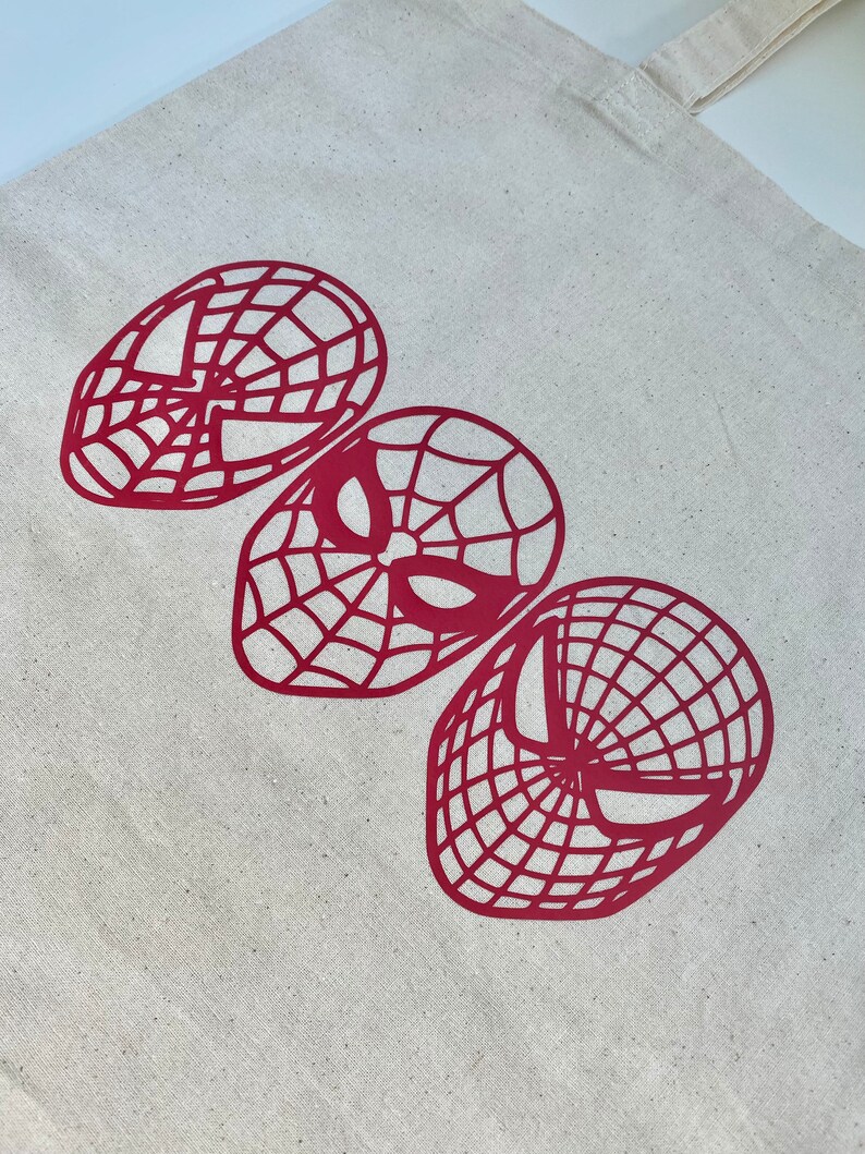 Puede incluir: Un bolso tote de lona beige con tres dise&ntilde;os de m&aacute;scaras rojas de Spider-Man. Las m&aacute;scaras presentan diferentes estilos, con patrones de telara&ntilde;a y formas de ojos. El bolso tiene una forma rectangular simple con dos asas.