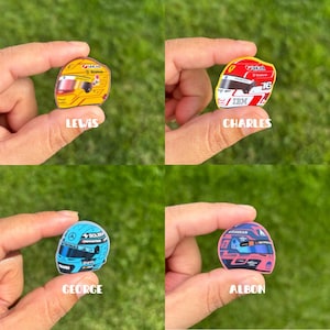 Pode incluir: Quatro pequenos adesivos em formato de capacetes de corrida. Um &eacute; amarelo com o texto LEWIS, um vermelho e branco com o texto CHARLES, um azul claro com o texto GEORGE e um rosa e azul com o texto ALBON, cada um segurado por dedos.