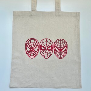 Puede incluir: Una bolsa de mano de lona beige con tres dise&ntilde;os de m&aacute;scaras de Spider-Man rojas. Las m&aacute;scaras est&aacute;n dispuestas horizontalmente en la parte delantera de la bolsa, cada una con un patr&oacute;n de telara&ntilde;a &uacute;nico. La bolsa tiene dos asas beige a juego.
