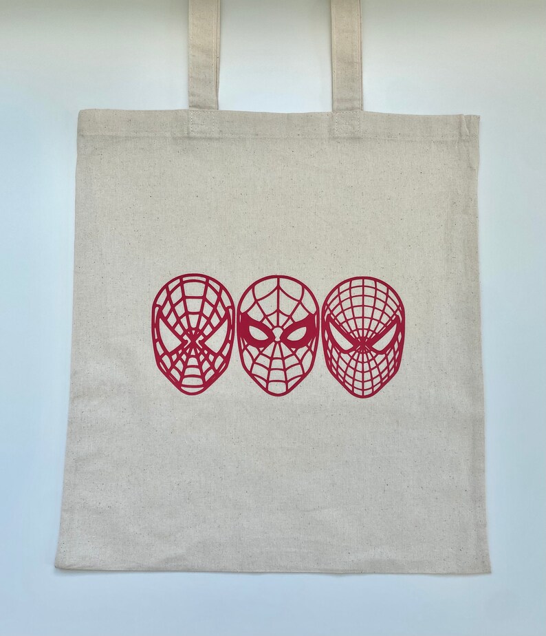 Puede incluir: Una bolsa de mano de lona beige con tres dise&ntilde;os de m&aacute;scaras de Spider-Man rojas. Las m&aacute;scaras est&aacute;n dispuestas horizontalmente en la parte delantera de la bolsa, cada una con un patr&oacute;n de telara&ntilde;a &uacute;nico. La bolsa tiene dos asas beige a juego.