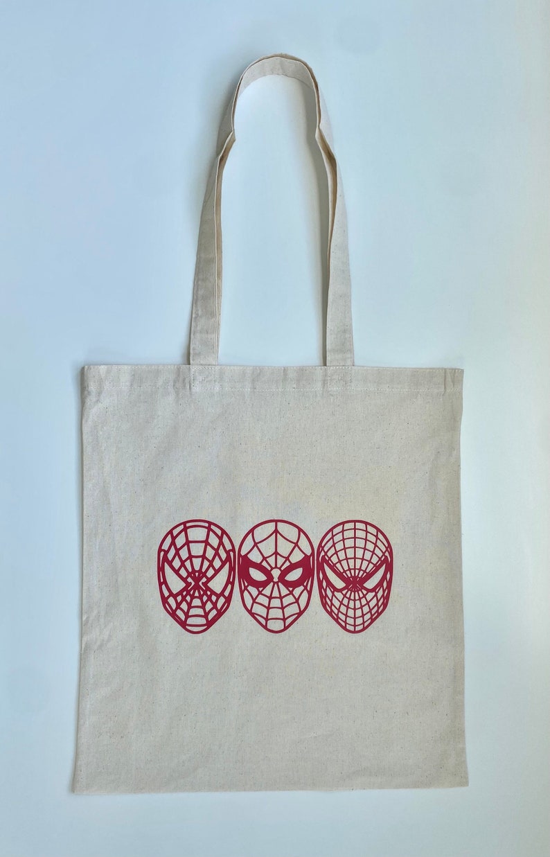 Puede incluir: Un bolso tote de lona beige con tres dise&ntilde;os de m&aacute;scaras rojas de Spider-Man. El bolso tiene dos asas y est&aacute; hecho de una tela resistente. Las m&aacute;scaras de Spider-Man est&aacute;n en fila.