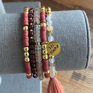 May include: A stack of five bracelets with a variety of beads and accents. The bracelets feature gold, maroon, clear, and light-colored beads. A gold charm with the text "S&L Stackz" hangs from one bracelet, and a maroon tassel hangs from another.