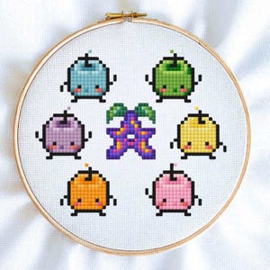 May include: Cross-stitch embroidery hoop with a colorful design of cartoon fruit characters. The fruit characters are a purple plum, a green apple, a yellow lemon, a pink strawberry, an orange peach, and a blue blueberry. The characters are all smiling and have arms and legs. In the center of the hoop is a purple flower with five petals.