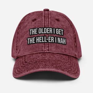 May include: A maroon baseball cap with a distressed, washed-out texture. The front features white embroidered text that reads THE OLDER I GET THE HELL-ER I NAH in two lines, with each line enclosed in a black rectangular border.