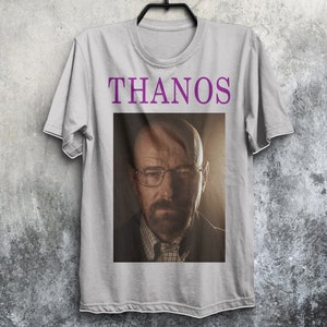 Op de afbeelding: Een grijs T-shirt met het woord "THANOS" in paars op de voorkant. Het shirt heeft een afbeelding van een man met een baard en bril.