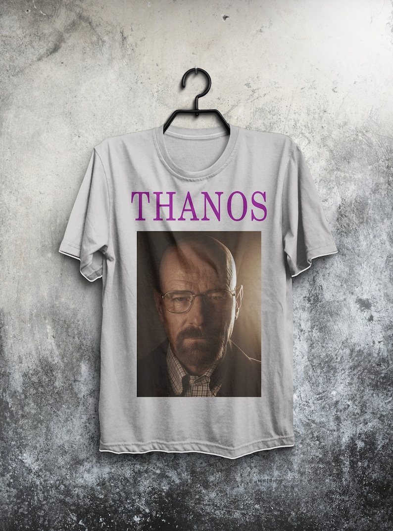 Op de afbeelding: Een grijs T-shirt met het woord "THANOS" in paars op de voorkant. Het shirt heeft een afbeelding van een man met een baard en bril.