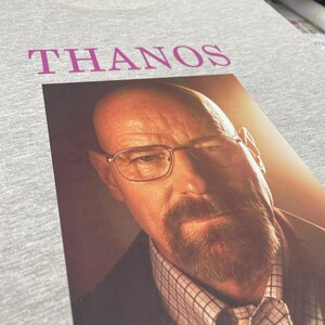 Op de afbeelding: Een grijs T-shirt met het woord "THANOS" in roze op de voorkant. Onder het woord is een foto van een man met een baard en bril gedrukt.