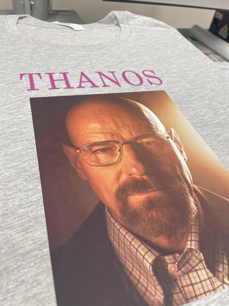 Op de afbeelding: Een grijs T-shirt met het woord "THANOS" in roze op de voorkant. Onder het woord is een foto van een man met een baard en bril gedrukt.