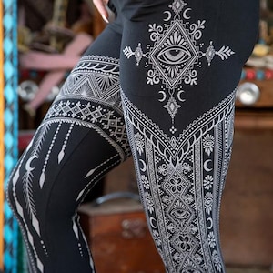 May include: Black leggings with intricate white geometric patterns. The design includes eye motifs, crescent moons, and diamond shapes. The leggings have a high waist and a pocket. The overall style is bohemian and spiritual.