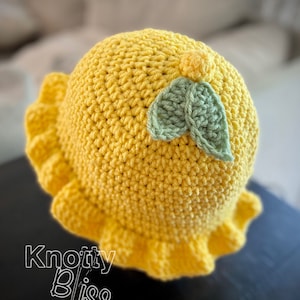 Pu&ograve; includere: Un cappello all'uncinetto giallo con un design a foglia di uncinetto verde. Il cappello ha un bordo arricciato e il testo "Knotty Bliss" &egrave; visibile nell'angolo in basso a sinistra dell'immagine.