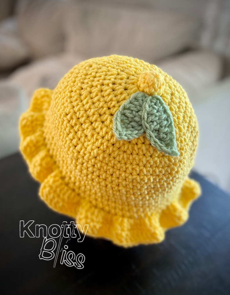 Pu&ograve; includere: Un cappello all'uncinetto giallo con un design a foglia di uncinetto verde. Il cappello ha un bordo arricciato e il testo "Knotty Bliss" &egrave; visibile nell'angolo in basso a sinistra dell'immagine.