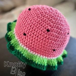 Pu&ograve; includere: Un cappello a uncinetto a forma di anguria con un corpo rosa, strisce verdi e bianche e semi neri. Il cappello ha un bordo arricciato.