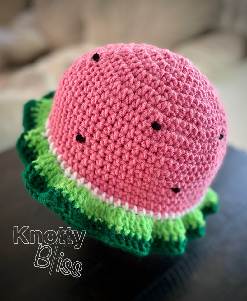 Pu&ograve; includere: Un cappello a uncinetto a forma di anguria con un corpo rosa, strisce verdi e bianche e semi neri. Il cappello ha un bordo arricciato.