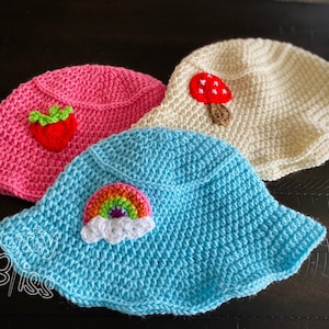 May include: Three crocheted sun hats in pink, white, and blue. The pink hat has a strawberry applique, the white hat has a mushroom applique, and the blue hat has a rainbow applique.