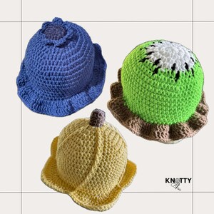 Pu&ograve; includere: Tre cappelli all'uncinetto in diversi colori e design. Un cappello blu con un fiore in cima, un cappello verde con un design a frutto di kiwi e un cappello giallo con un design a banana.