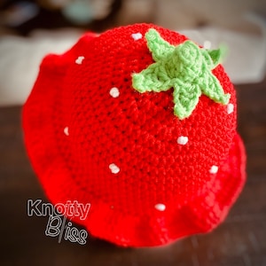 Pu&ograve; includere: Un cappello di fragola rosso all'uncinetto con una foglia verde all'uncinetto in cima. Il cappello ha dei puntini bianchi in tutto.