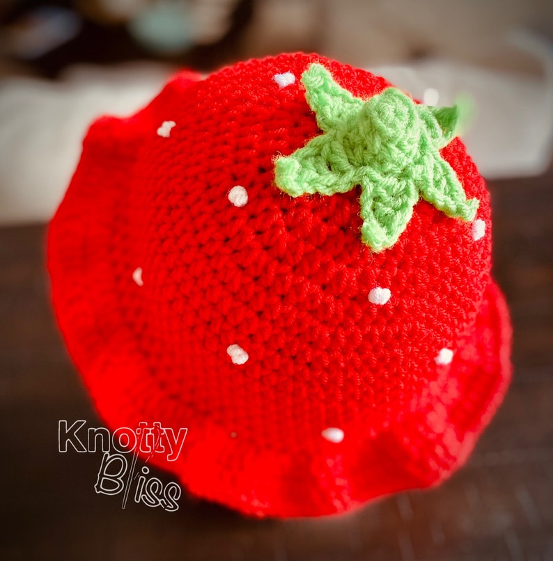 Pu&ograve; includere: Un cappello di fragola rosso all'uncinetto con una foglia verde all'uncinetto in cima. Il cappello ha dei puntini bianchi in tutto.