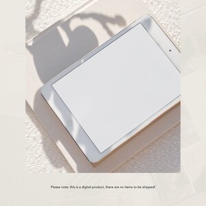 May include: A white tablet computer with a blank screen is lying on a beige surface. The tablet is surrounded by shadows from the sun.